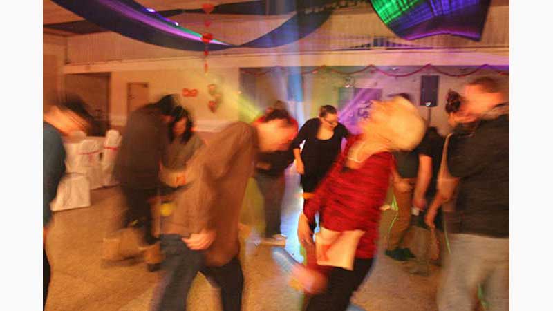  Fundraiser DJ, DJ Burlington, Blurred action photo of people dancing and lady in forefront laughing whole heartedly. Colored light beams coming from behind the dancers and shining over their heads. Taken in Burlington Ontario
